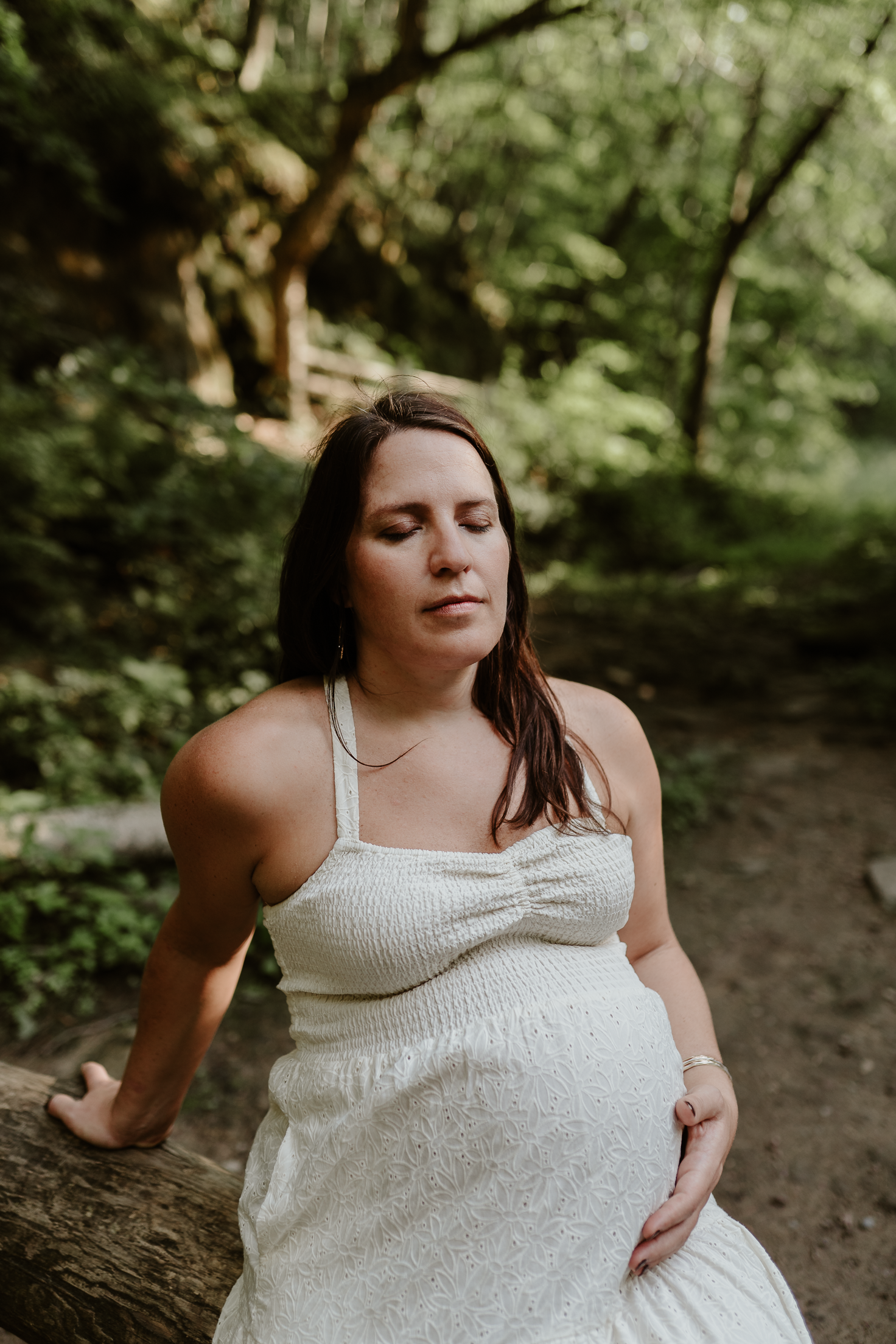 Pregnant mother in a white, eyelit dress sitting on a log in a creek with her eyes closed.