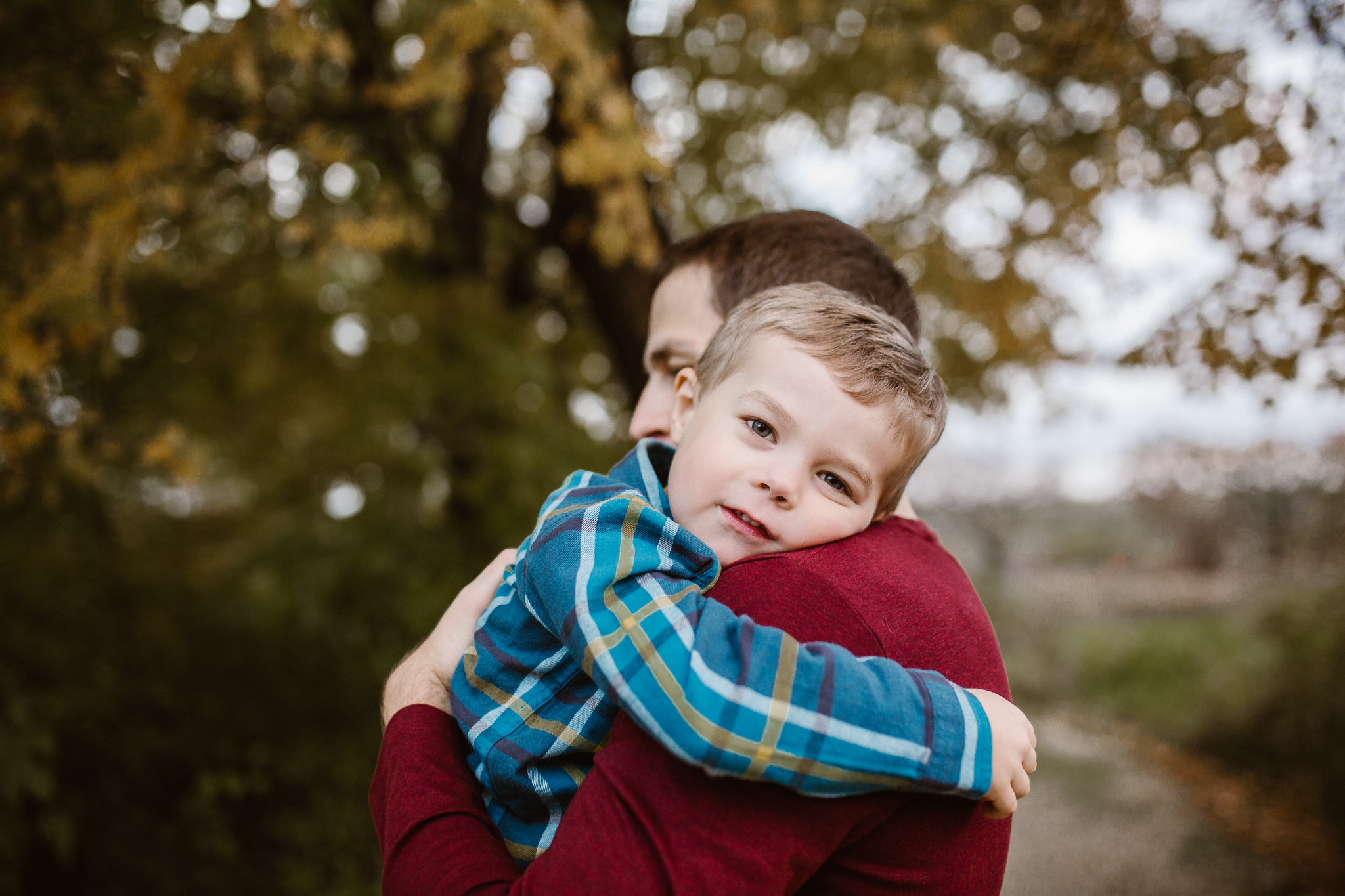 Son resting his head on his father's shoulder in the fall trees.