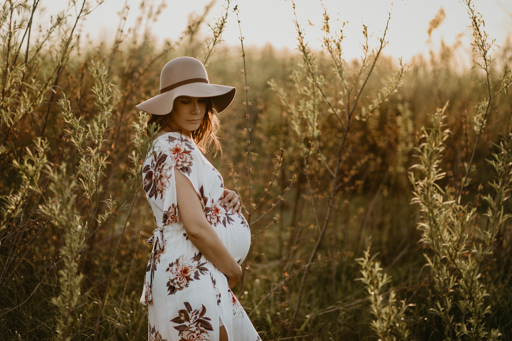 Expecting woman in a sun hat holding her belly in a golden field of grasses.