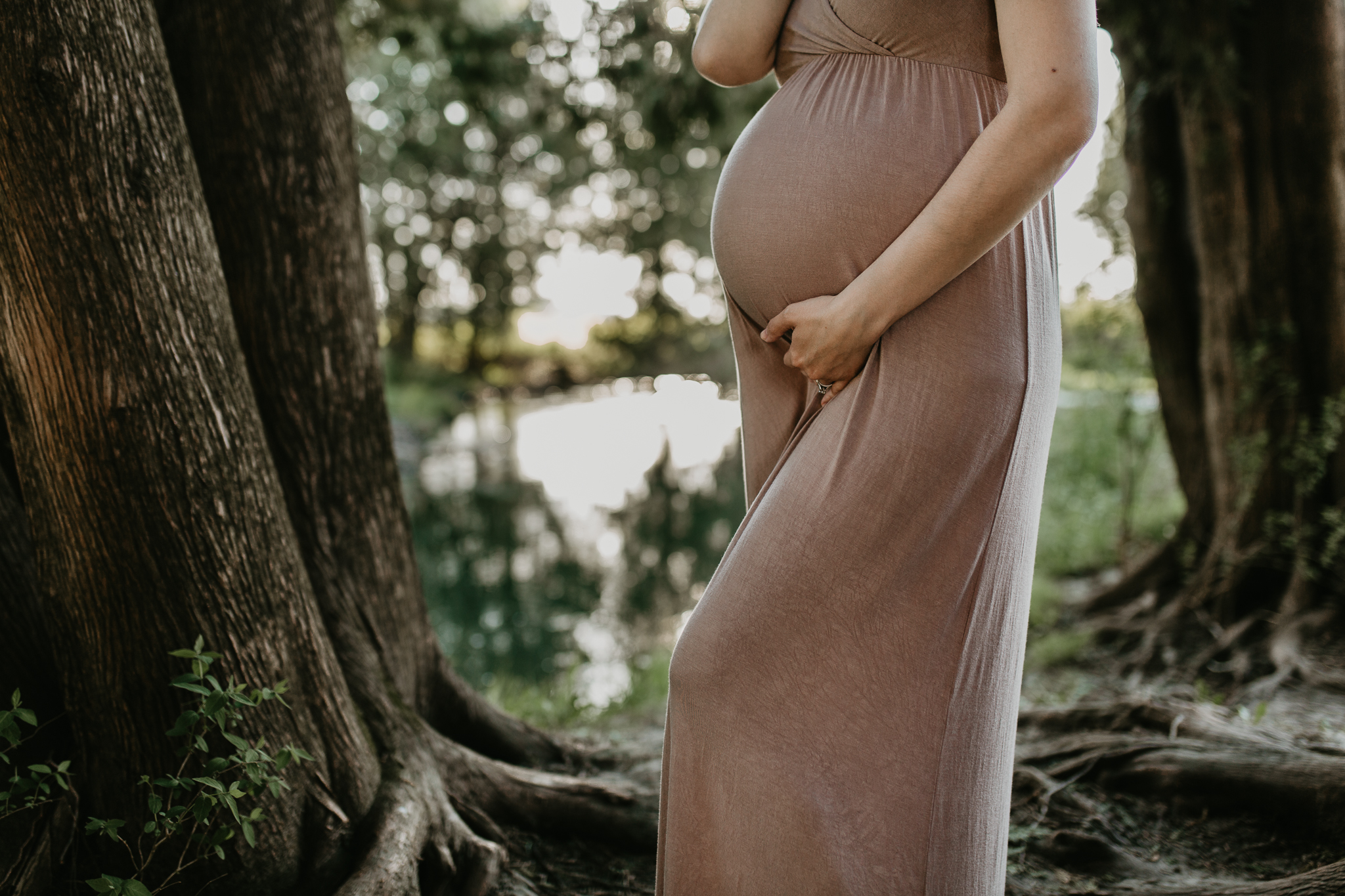 Pregnant woman in a long maternity dress in the woods in front of a stream.