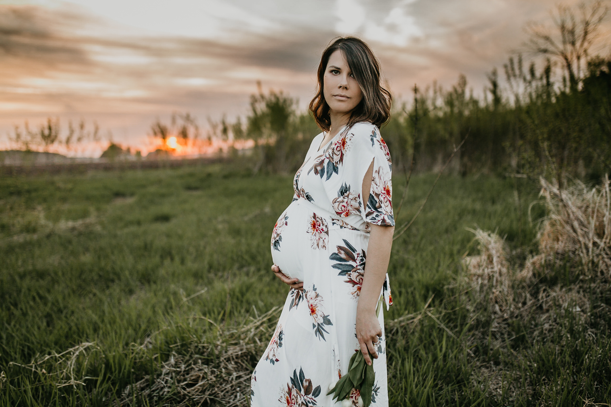 Pregnant mother in a grass field at golden hour holding the bottom of her belly and tulips.