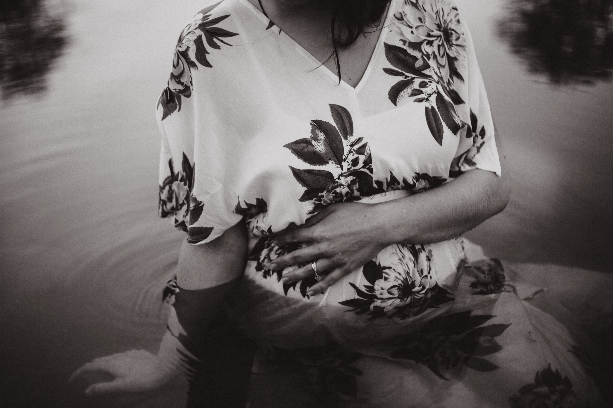 Expecing mother in a white floral dress sitting in the water holding her pregnant belly.