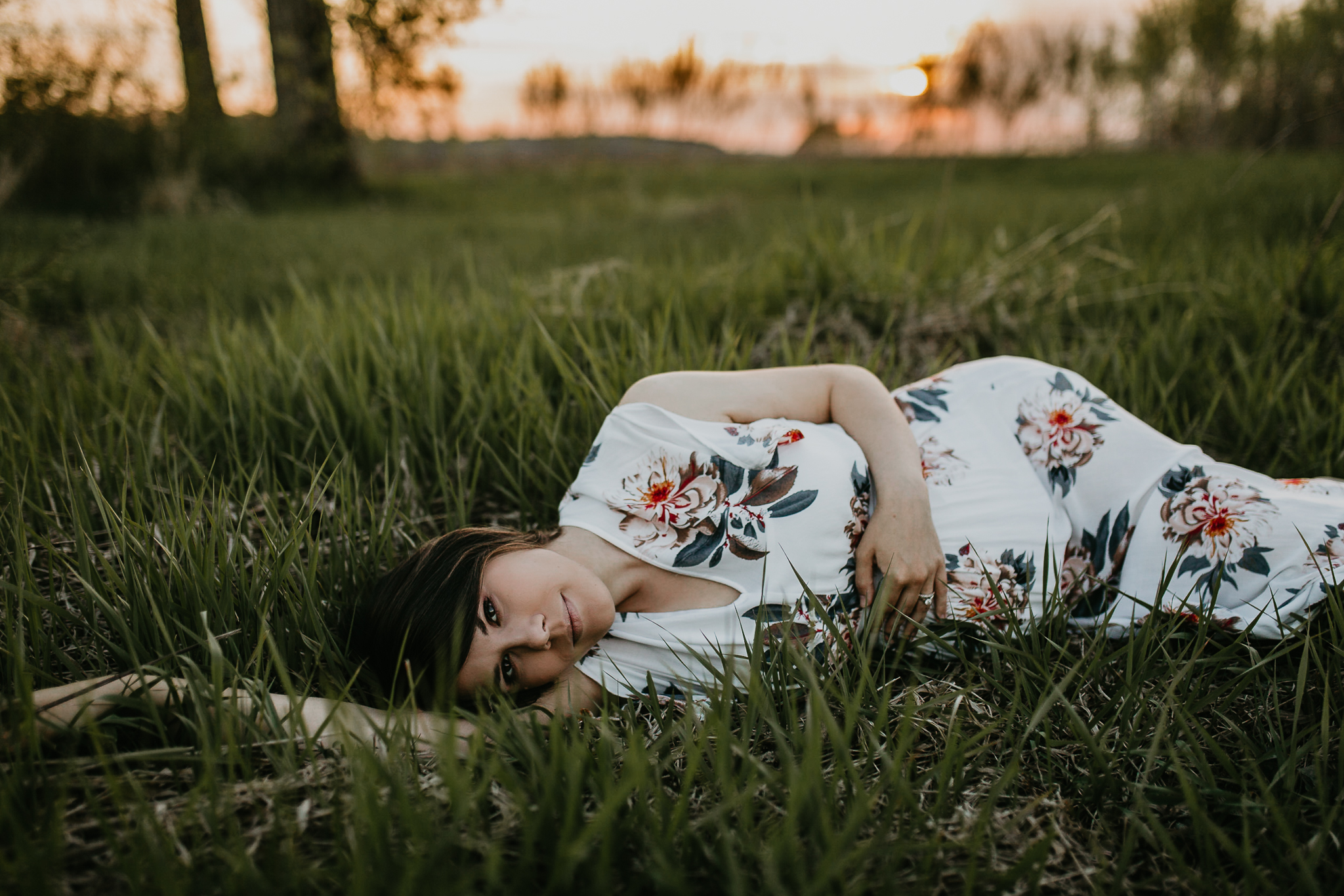 Expecting woman in a floral maxi dress laying down in a grass field during sunset.