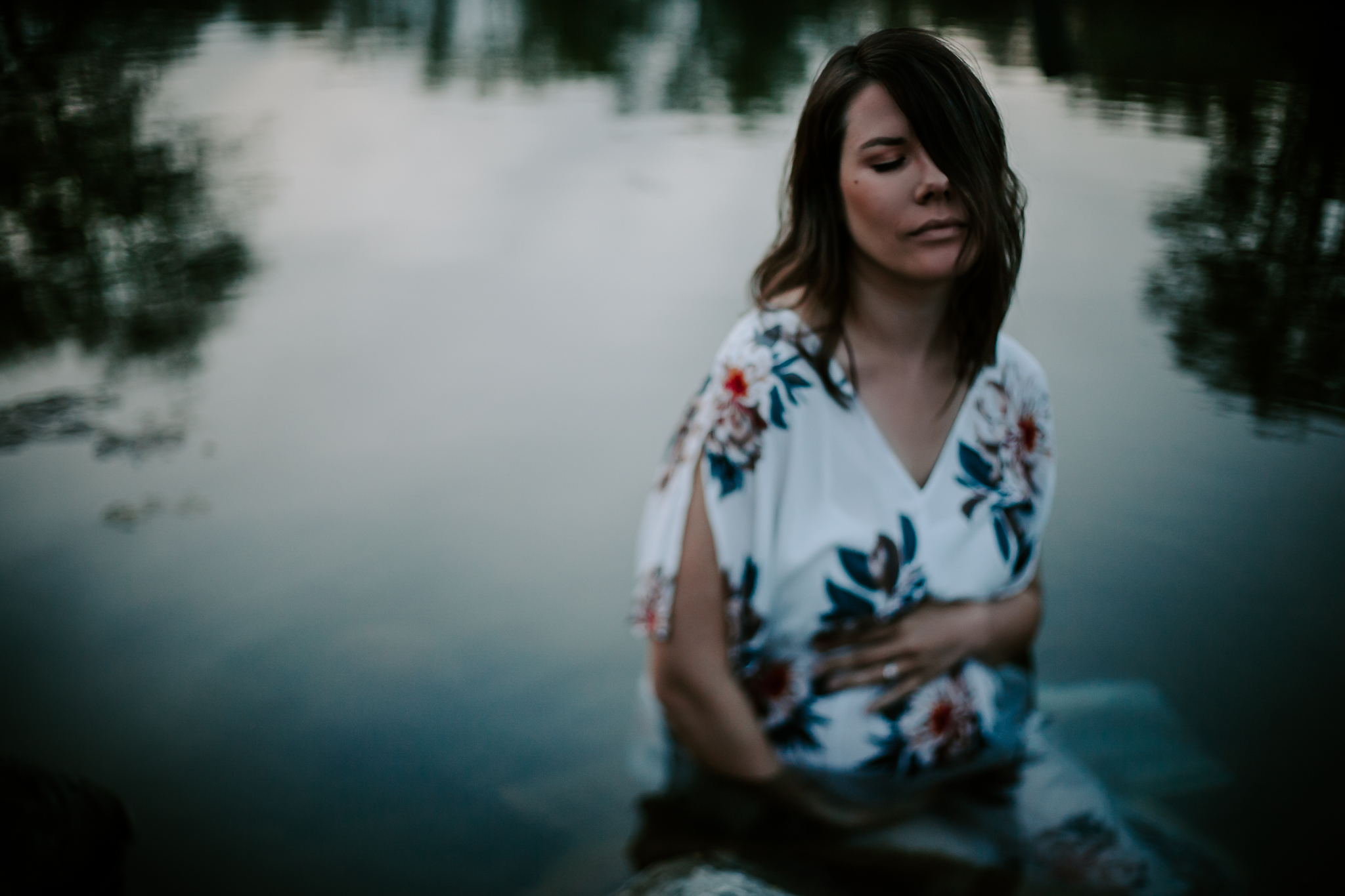 Expecing mother in a white floral dress serenly sitting in pond holding her belly at blue hour.