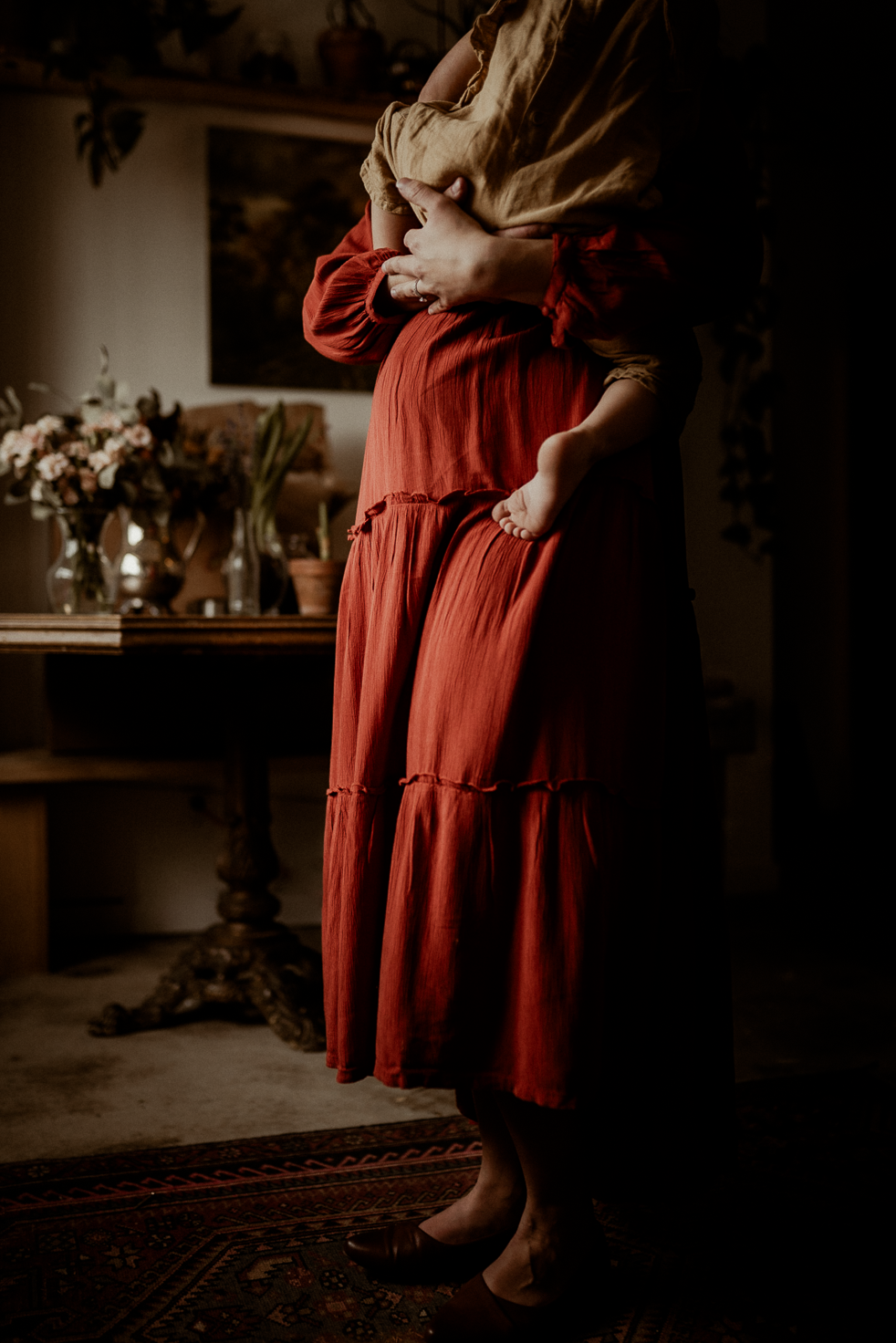 Pregnant mother in a rust dress holing her young daughter up on her belly at a coffee shop.