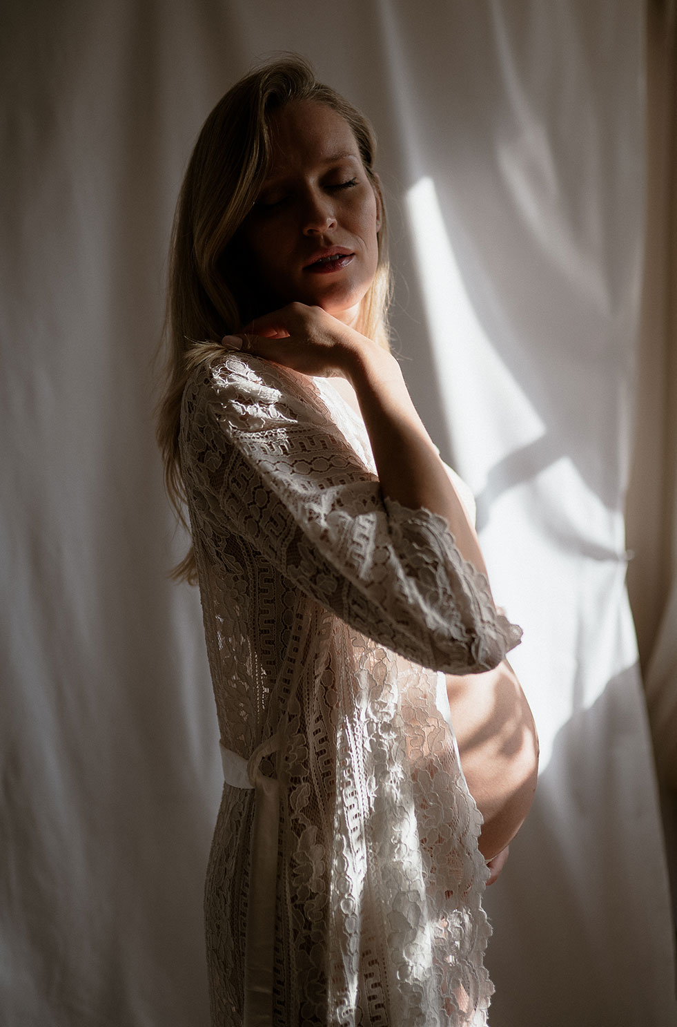 Pregnant woman in a white lace robe with shadows on her belly.