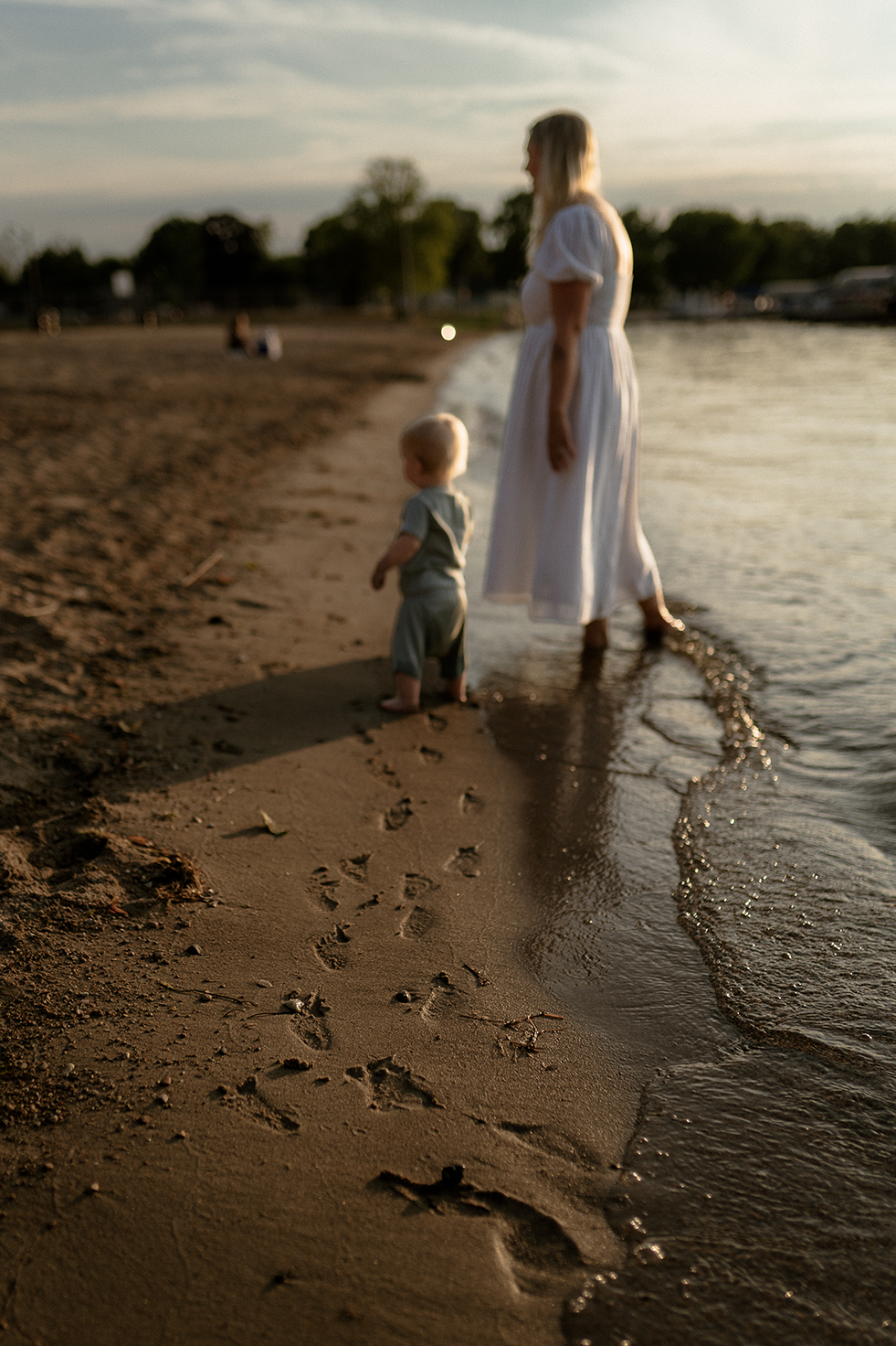 Mother in a long white dress walking with her baby son along the beach leaving footprints behind them in the sand.