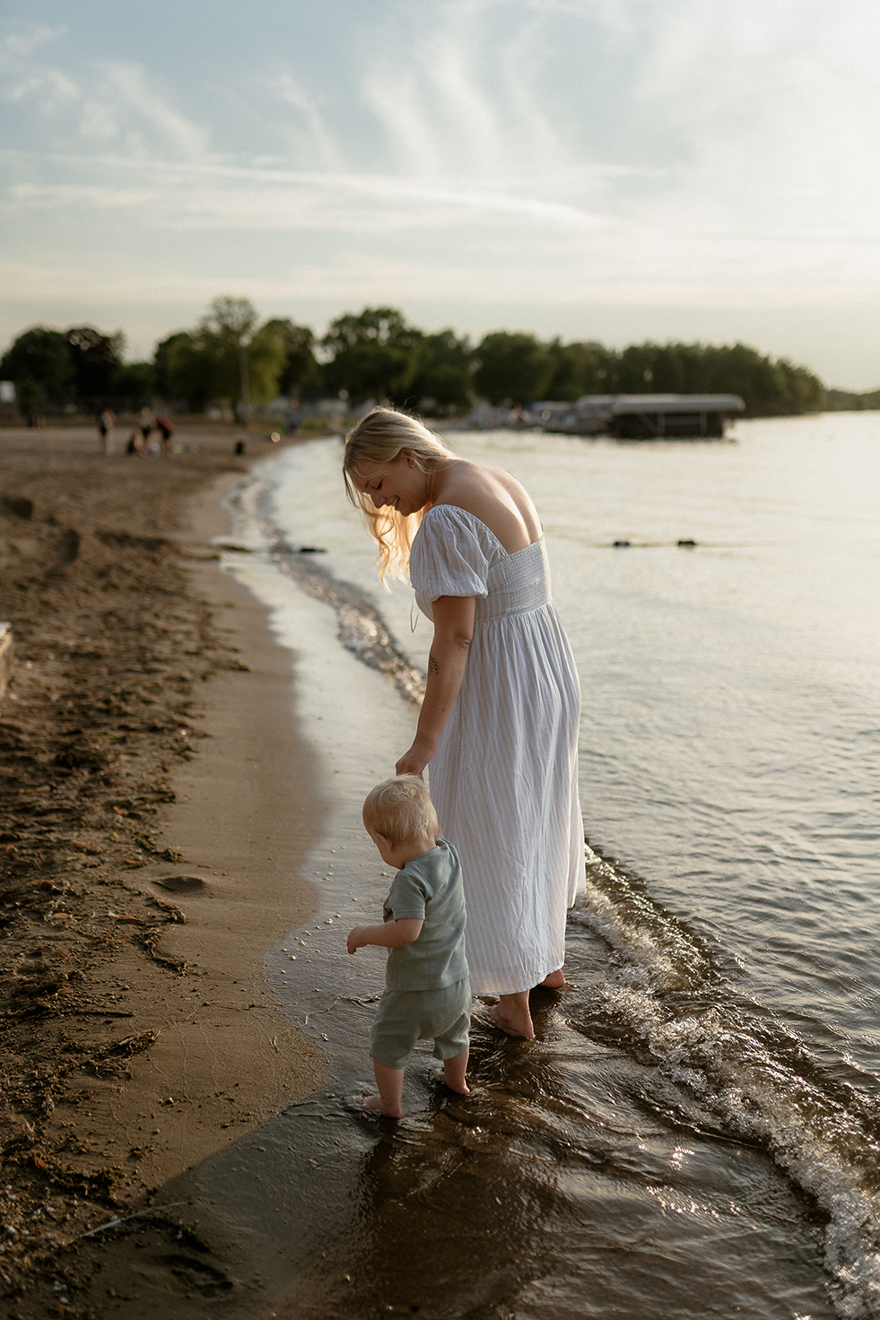 Mother in a white dress holding her baby sons hand as they walk and explore the beach lakefront together.